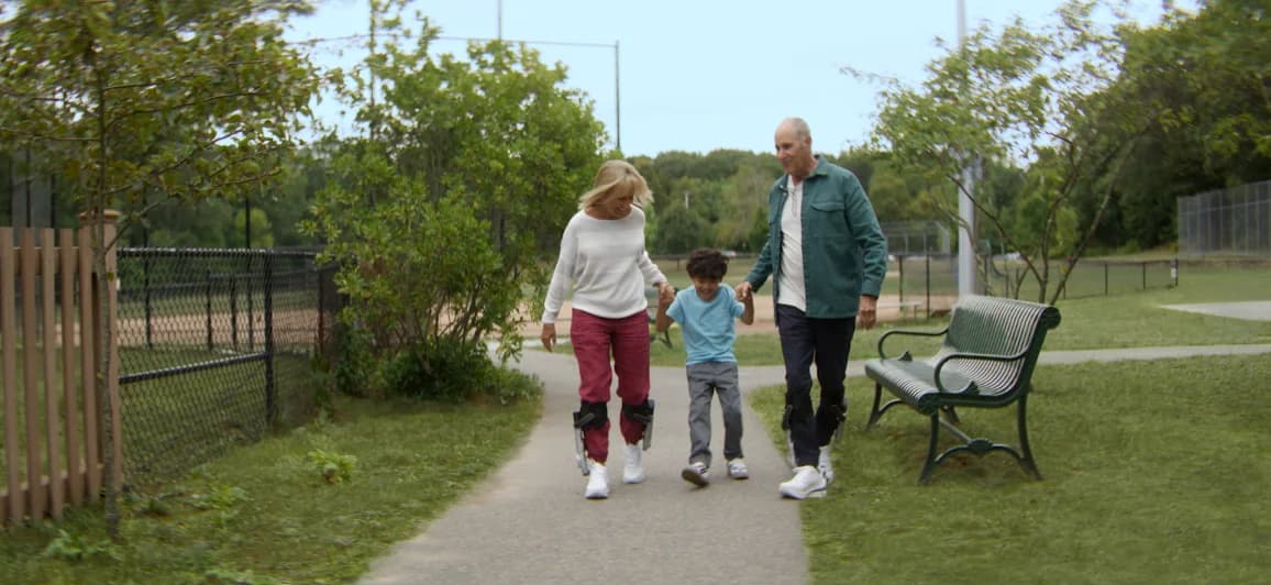 Two adults wearing Dephy Sidekick walk with a child along a park path.