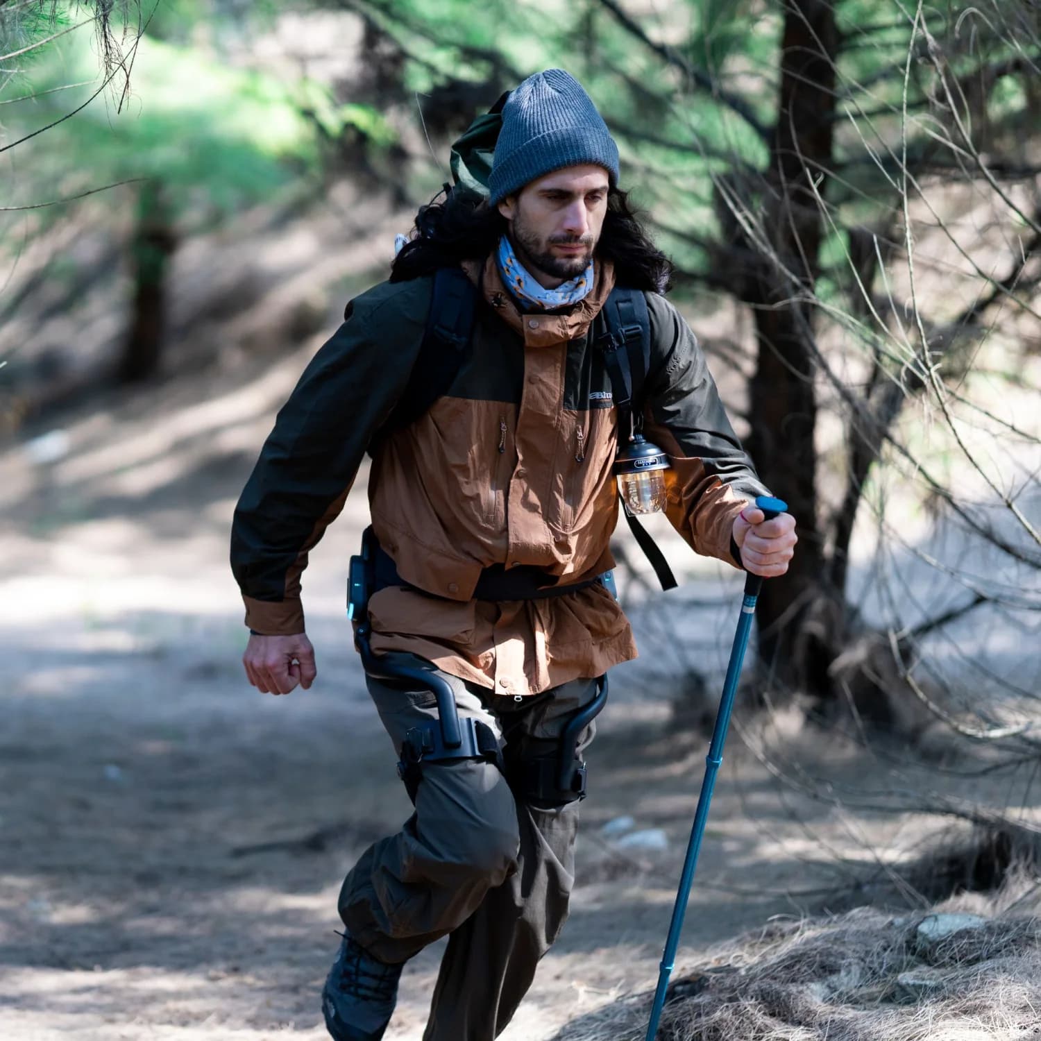 A hiker wearing a DNSYS X1 walks on a wooded trail with hiking poles.